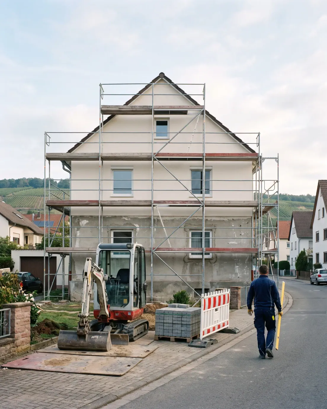 Einfamilienhaus in Sanierung: eingerüstete Fassade, halb neu verputzt, Minibagger im Vorgarten